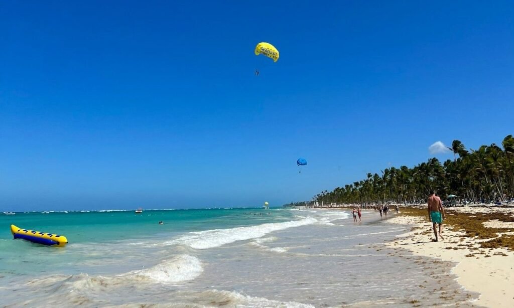 Punta Cana beach with turquoise water, palm trees, parasailing, and people walking along the white sand shoreline in the Dominican Republic.